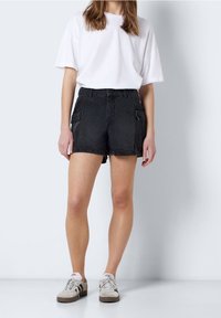 Schwarze Jeansshorts mit hoher Taille, seitlichen Cargo-Taschen und einer strukturierten Oberfläche. Kombiniert mit einem schlichten weißen oversized T-Shirt und beigen Sneakers.