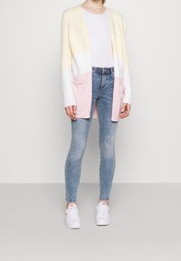 Larga chaqueta en amarillo pastel, blanco y rosa con un frente abierto, dos bolsillos, combinada con jeans ajustados azul claro y zapatillas blancas.