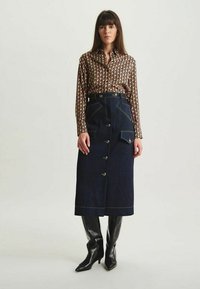 Chemise à boutons en tissu à motifs, associée à une jupe en denim foncé avec des poches et des boutons métalliques, stylée avec des bottes montantes.