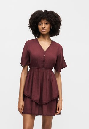 Γυναίκα με κατσαρά μαλλιά, που φοράει ένα μπορντό φόρεμα με κοντά μανίκια, V-λαιμόκοψη και φούστα με στρώσεις, στέκεται μπροστά σε έναν απλό φωτεινό φόντο.