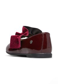 Scarpa borgogna in vernice con punta arrotondata e suola nera, dotata di un ampio fiocco texturizzato in color marrone scuro sul lato.