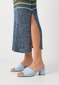 Femme portant une jupe tricotée bleu et vert avec une fente haute sur le côté et des mules à talon bloc ouvertes bleu clair sur un sol blanc.