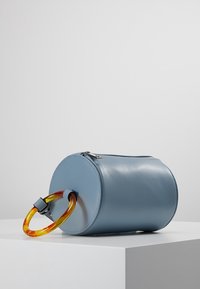 Borsa rotonda in pelle blu con chiusura a zip e manico in acrilico arancione traslucido. Texture liscia, design minimalista e senza decorazioni aggiuntive.