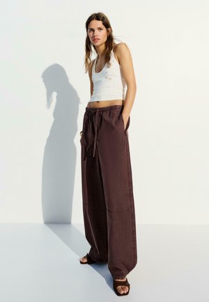 Kvinde står op ad hvid væg iført hvidt tanktop, løse brune bukser og brune sandaler med hænderne i lommerne.