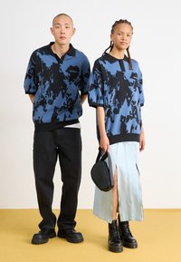 Δύο μοντέλα φορούν ένα oversized πλεκτό πόλο σε μπλε και μαύρο σχέδιο και μια ανοιχτόχρωμη τζιν φούστα με πλευρικούς φερμουάρ. Περιλαμβάνονται μαύρες στρατιωτικές μπότες και μια μικρή τσάντα.