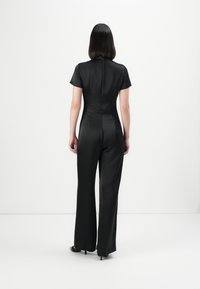 Woman with straight black hair wearing a black short-sleeve jumpsuit and black heels standing with back to camera on white background.