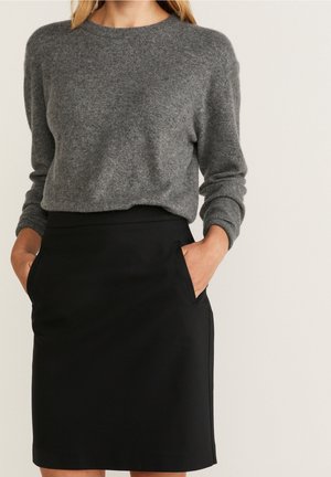 Kvinde iført en grå langærmet sweater og en sort knælængde nederdel med hænderne i lommerne mod en ensfarvet baggrund.