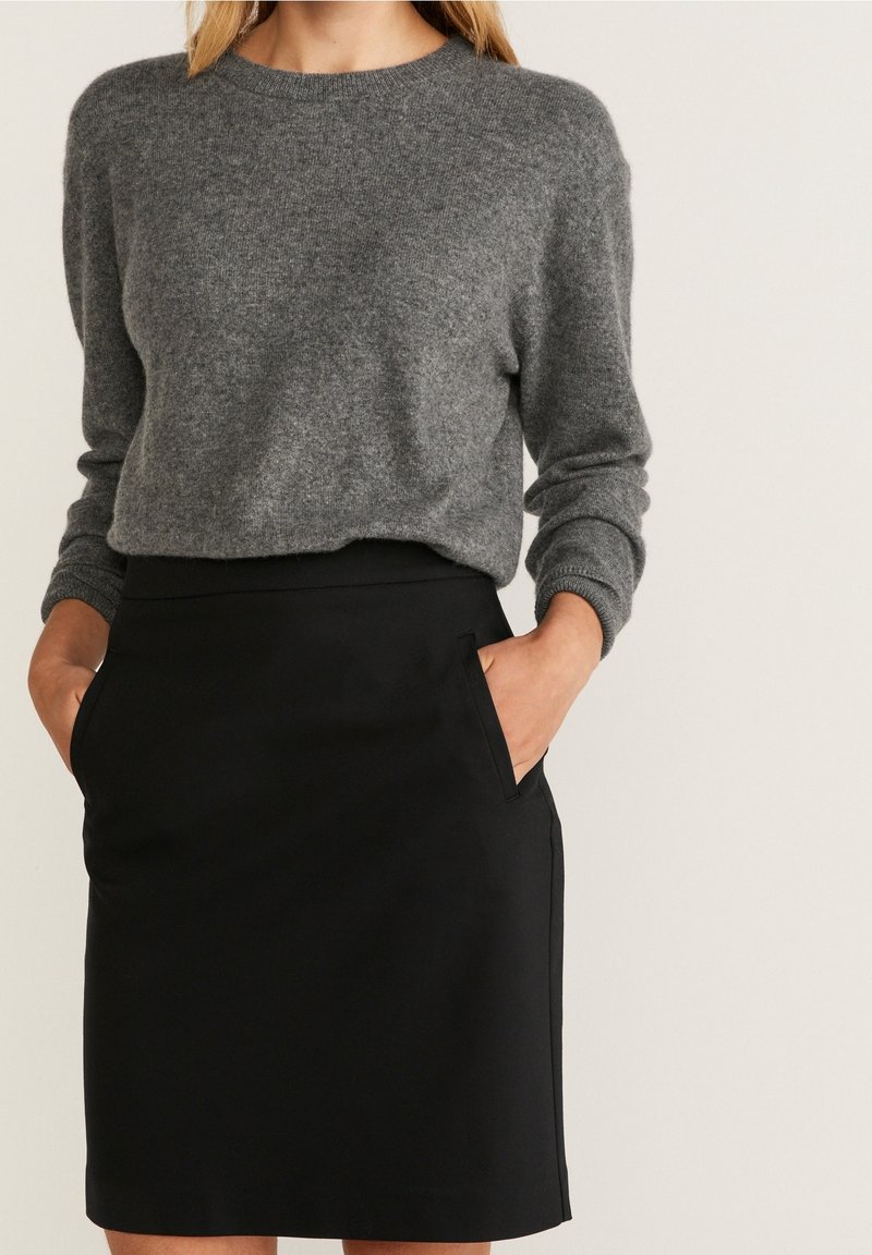 Femme portant un pull gris à manches longues et une jupe noire mi-longue, les mains dans les poches, sur un fond uni.