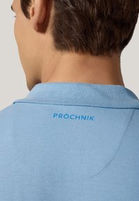 Jasnoniebieska koszulka polo z teksturowanego dzianiny, z kołnierzykiem i nazwą marki "PRÓCHNIK" w niebieskim kolorze na plecach.