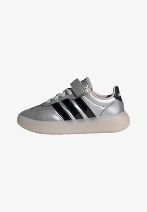 Zapatillas de cuero plateado con franjas negras brillantes, puntera redonda y una tira de velcro. Cuentan con una suela de goma translúcida y texturizada.