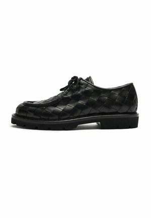 Zapato negro de cuero con cordones y patrón trenzado, suela de goma gruesa y punta redonda, mostrado en perfil lateral sobre fondo blanco.