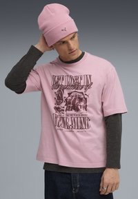 T-shirt rosa a maniche corte con design grafico di testo. Sovrapposto a un top lungo scuro. La modella indossa un berretto di maglia rosa abbinato. Jeans in denim.