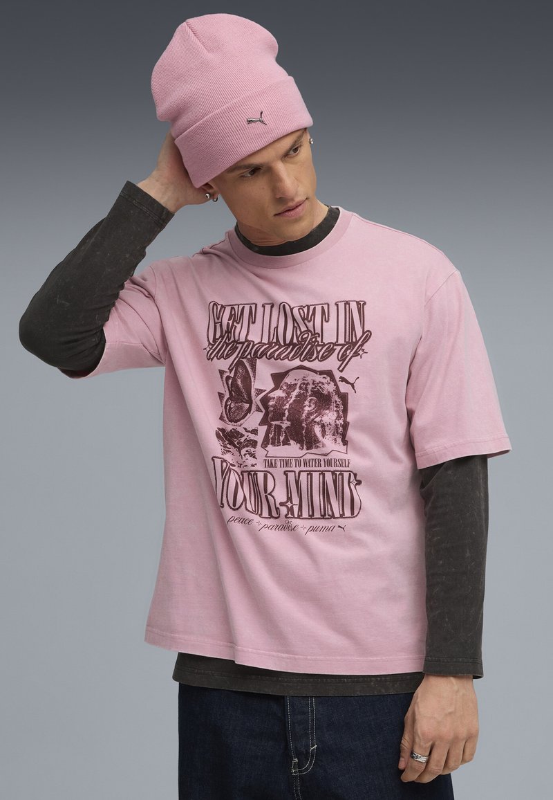 Camiseta rosa de manga corta con diseño gráfico de texto. Superpuesta a una camiseta de manga larga oscura. El modelo lleva un gorro de punto rosa a juego. Jeans de mezclilla.