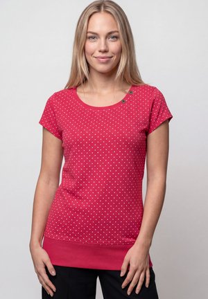 Young woman with straight blonde hair wears a red, short-sleeve, polka dot top with black pants against a plain background.