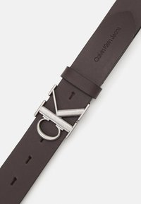 Barna bőr öv, ezüstszínű fémcsattal, amelyen a jellegzetes Calvin Klein logó található, valamint több állítható lyukkal.