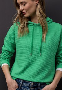 Femme portant un sweat à capuche vert texturé et un jean bleu, debout devant un fond gris foncé, regardant vers le bas et sur le côté, les mains dans les poches.