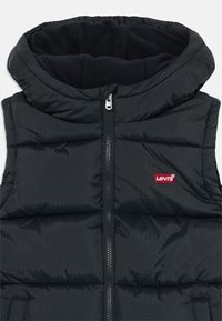 Juodas siūtas be rankovių pūkuotas liemenė su aukštu apykakle, centriniu užtrauktuku ir raudonu Levi's logotipu kairėje krūtinės pusėje.