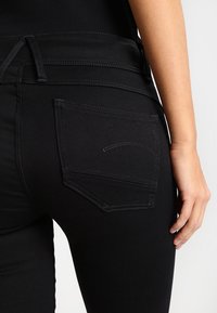 Jeans de mezclilla negra con un diseño ajustado, que presentan un único bolsillo trasero con un detalle de costura curva. Textura suave y estilo clásico.