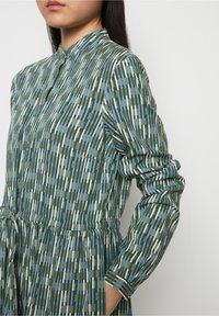 Abito verde a fantasia con righe verticali in blu e crema, colletto alla cinese, chiusura con bottoni e vita arricciata con tasche.