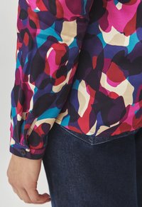 Blusa colorata con un audace motivo astratto in tonalità di rosa, blu e colori scuri. Maniche lunghe con polsini a bottone e una texture liscia.