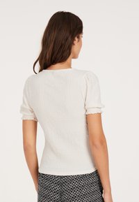 Hvid kortærmet top med et tekstureret, ribbet design, der har en rund halsudskæring og samlede ærmer, parret med mønstrede sorte bukser.