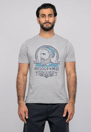 Homme aux cheveux courts et foncés et barbe, portant un t-shirt gris clair avec un motif nautique bleu et noir, et un pantalon foncé, devant un fond uni.