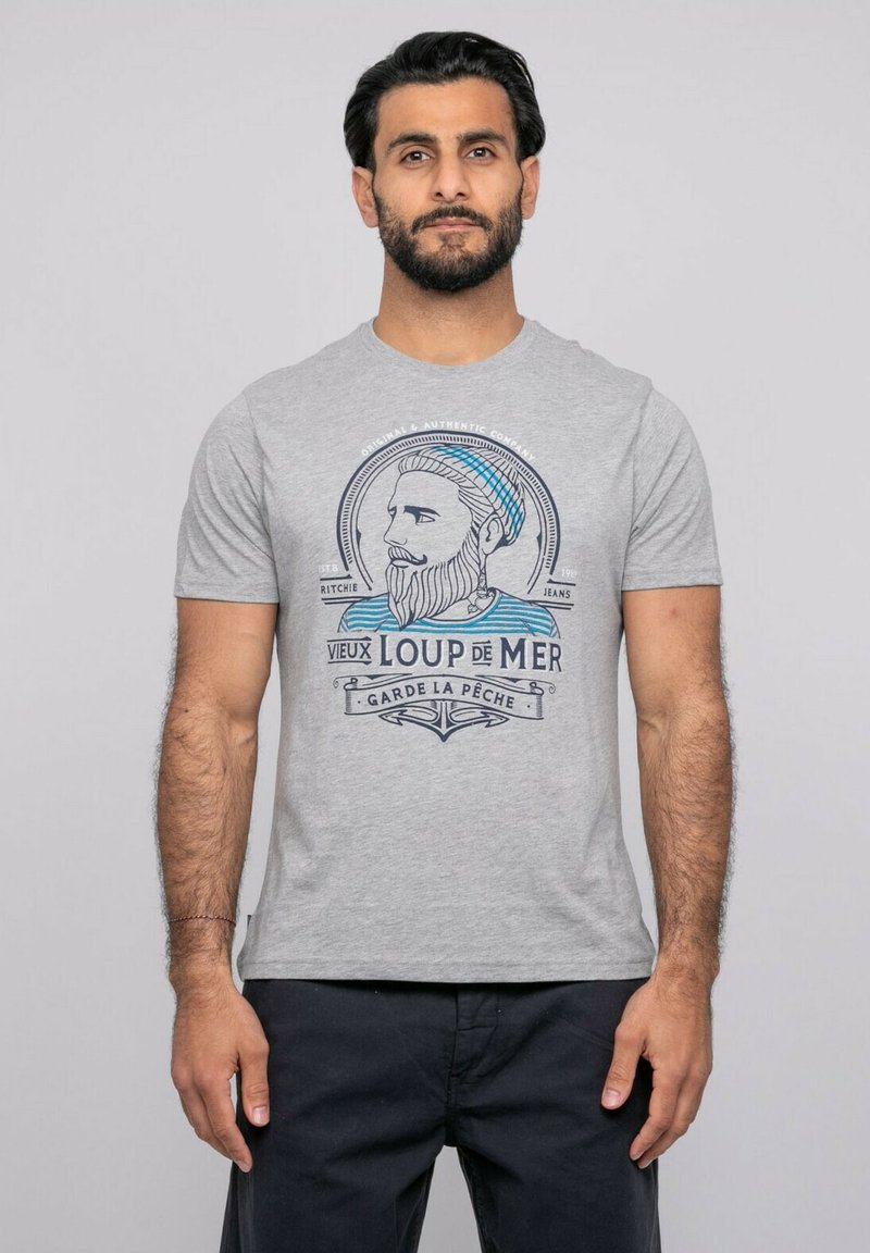 Homme aux cheveux courts et foncés et barbe, portant un t-shirt gris clair avec un motif nautique bleu et noir, et un pantalon foncé, devant un fond uni.