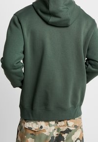 Grön sweatshirt med huva, med en mjuk textur. Parat med byxor i kamouflagemönster, som visar jordnära färger och en avslappnad passform.