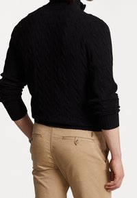 Schwarzer, kabelgestrickter Pullover mit hohem Kragen und gerippten Bündchen, kombiniert mit khakifarbenen Hosen, die über Gesäßtaschen und ein kleines Logo-Detail verfügen.