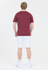 Mand der står med ryggen til iført en bordeaux farvet T-shirt med kort ærme, hvide shorts, hvide sokker og hvide sneakers med guldaccent.