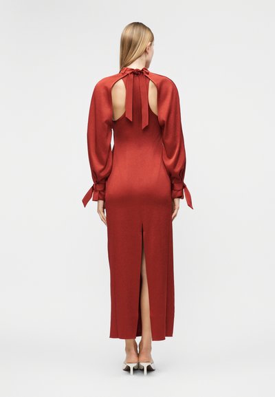 Γυναίκα που φορά ένα μακρύ κόκκινο φόρεμα με φουσκωτά μανίκια, ανοιχτή πλάτη με κορδόνια στον λαιμό και τις μανσέτες, και σκίσιμο στο πίσω μέρος, στέκεται μπροστά σε απλό φόντο.