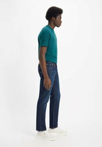T-shirt in cotone verde acqua, modello aderente, abbinato a jeans dritti blu scuro e sneakers bianche, vista laterale su uno sfondo uniforme.