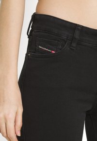Vaqueros de mezclilla negra con un diseño ajustado, bolsillos laterales y una pequeña etiqueta de marca con acentos rojos en el bolsillo delantero.