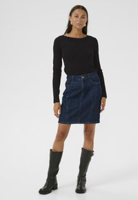 Denimrok in donkerblauw, met een getailleerd ontwerp, twee voorzakken en verticale naaddetails, gecombineerd met een zwarte top met lange mouwen en zwarte laarzen.