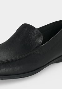 Mocassin en cuir noir à enfiler avec dessus texturé, couture visible et semelle en caoutchouc sur un fond clair uni.