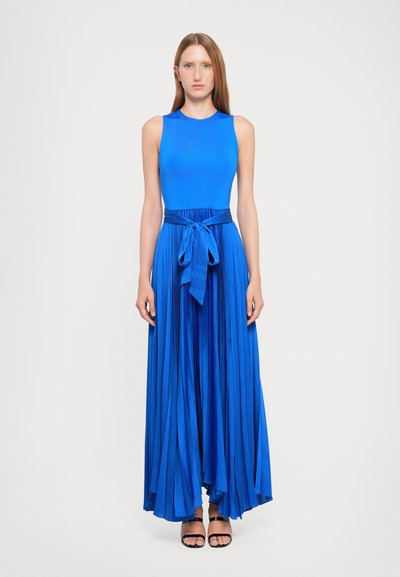 Top sans manches bleu associé à une jupe maxi plissée, mettant en avant une taille nouée. Texture lisse, couleur vibrante et silhouette fluide. Sandales noires à lanières.