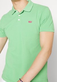 Man som bär en ljusgrön Levi's pikétröja med tre vita knappar och ett flätat armband på handleden.