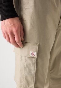 Beige cargo-housut, joissa on teksturoitu pinta, näkyvän läppätaskun ja punavalkoisen logopatchin sivussa.