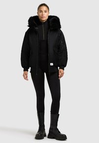 Giubbotto bomber nero con cappuccio in pelliccia sintetica, chiusura con zip, polsini elasticizzati, tasche laterali, abbinato a leggings neri e stivali militari con lacci.