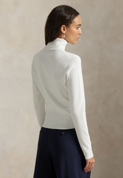 Mujer con cabello oscuro que lleva un suéter de cuello alto blanco y pantalones oscuros, de pie de perfil contra un fondo neutro.