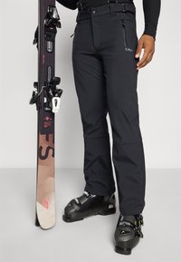 Pantaloni da sci neri con tasche frontali, indossati con stivali da sci neri e uno sci. I pantaloni presentano un design aderente e materiali impermeabili.