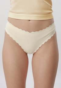 Femme portant des sous-vêtements couleur crème à bordures festonnées et un haut beige clair assorti, visibles de la taille jusqu'au milieu des cuisses, sur un fond uni.
