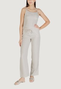 Jumpsuit beige chiaro con spalline sottili, vita elastica e gambe larghe, realizzato in tessuto morbido. Indossato con sandali chiari.