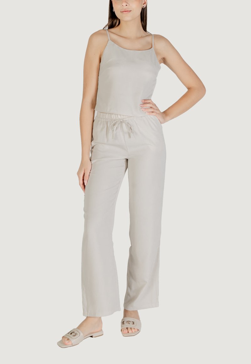 Jumpsuit beige chiaro con spalline sottili, vita elastica e gambe larghe, realizzato in tessuto morbido. Indossato con sandali chiari.