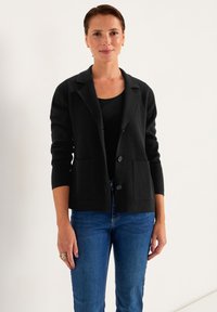 Sort strikket cardigan med klippet krave, to frontlommer og knaplukning. Båret over en sort bluse og parret med blå jeans.