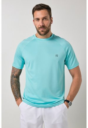 Mann mit tätowiertem linken Arm, trägt hellblaues kurzärmeliges Hemd und weiße Hose, Hände in den Taschen, neutrale Mimik vor grauem Hintergrund.