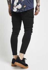 Jeans negros ajustados con detalles desgastados, combinados con zapatillas de ante negras con suela de goma. La parte superior tiene acentos florales en la parte posterior.