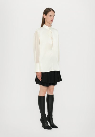 Silk blouse in off-white with a high collar and button detail, paired with a black pleated skirt and form-fitting black knee-high boots.