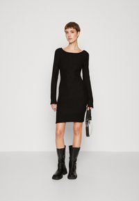 Black long-sleeve dress with a fitted silhouette, round neckline, and soft texture. Worn with black knee-high boots and a small bag.