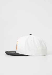 Hvid kasket med sort skygge og orange broderet detalje foran; lavet af stof med syninger og ventilationshuller.